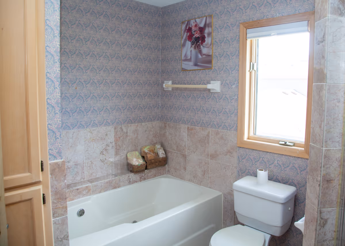 A bathroom with a white bathtub, a toilet with a roll of toilet paper on top, and a window with a wooden frame. The walls are decorated with patterned wallpaper and beige tiles. There is a wooden cabinet on the left side and a framed picture of flowers hanging above the bathtub. Two small baskets with toiletries are placed on the edge of the bathtub.