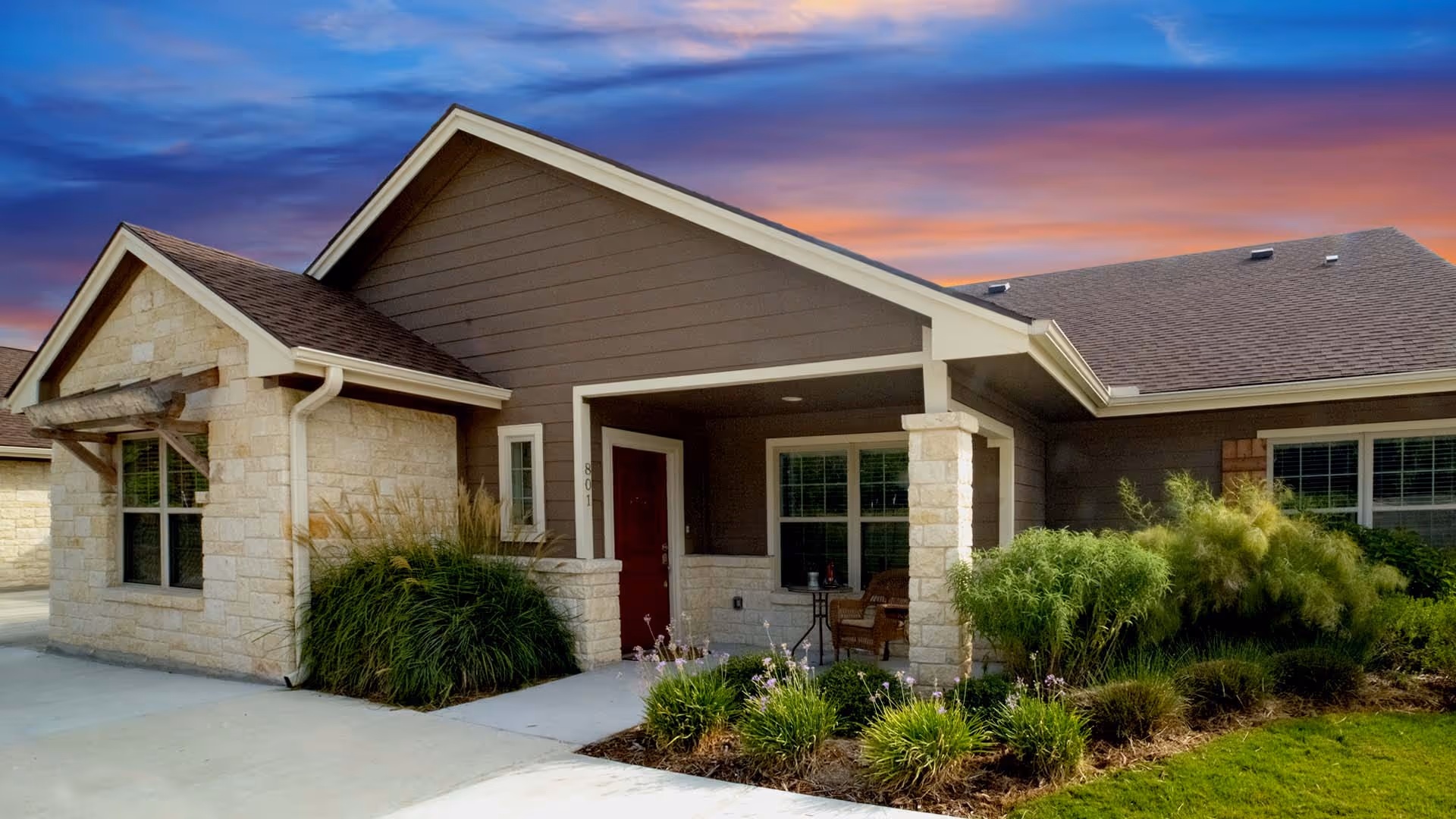 Single-story cottage-style building with a covered front porch, stone facade, and landscaped yard under a colorful sky.