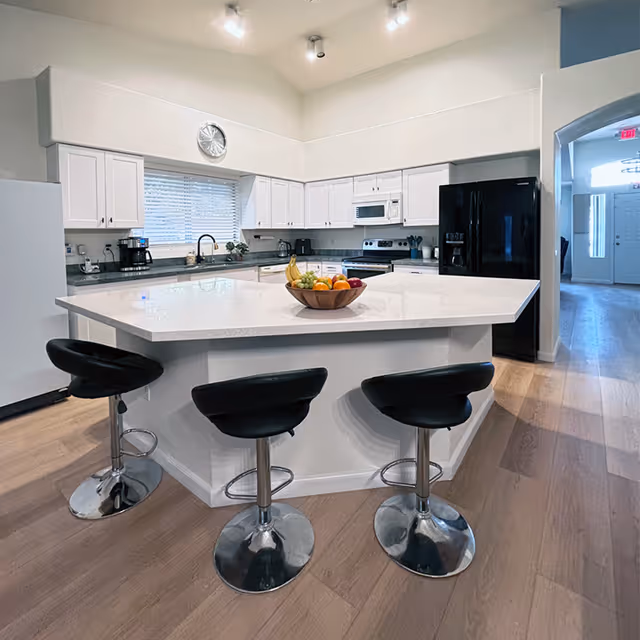 Modern kitchen with white cabinets, a large white island countertop with a bowl of fruit, three black bar stools, a black refrigerator, microwave, stove, coffee maker, and a window with blinds above the sink.