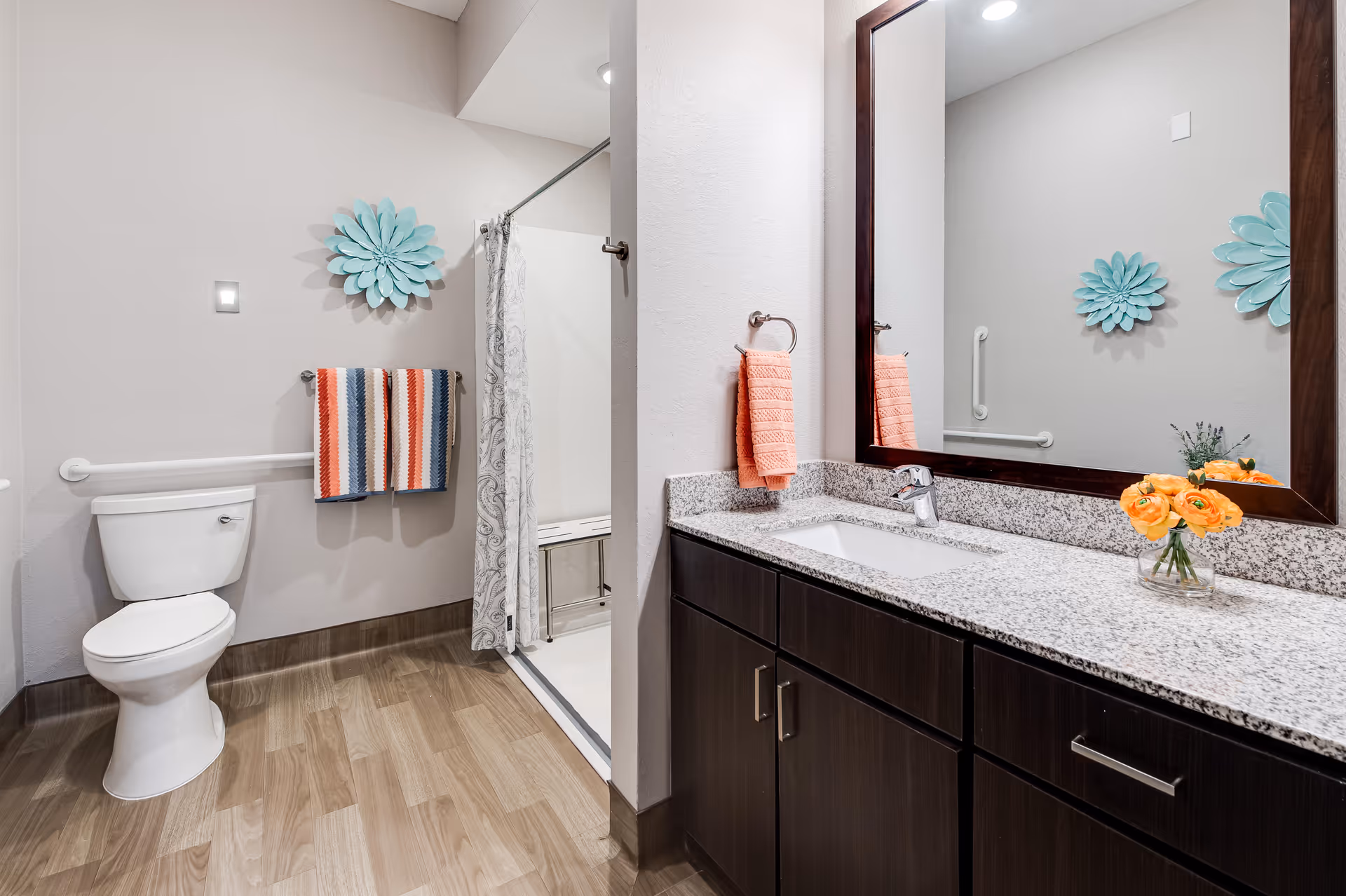 A modern bathroom with a white toilet, a walk-in shower with a patterned curtain, and a granite countertop with a sink. The walls are light-colored with a large mirror above the sink. There are colorful towels hanging on the wall and a small vase with orange flowers on the countertop. The floor has wood-like tiles and there is a decorative blue flower wall art above the toilet.