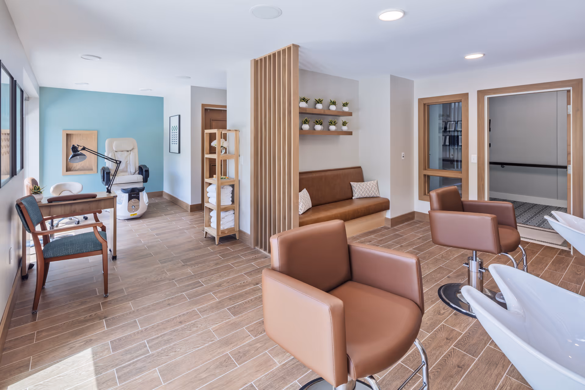 A bright and modern salon area in a senior living facility featuring two brown salon chairs in front of white washbasins, a brown cushioned bench with decorative pillows against the wall, wooden shelves with small potted plants, a wooden towel rack with folded white towels, a desk with a green chair and a lamp, and a white pedicure chair near a light blue accent wall.