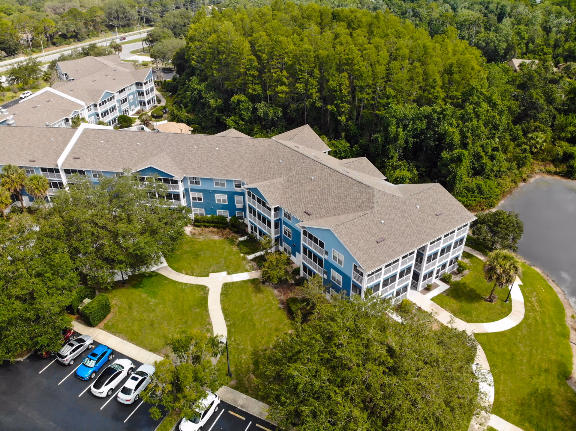 Aerial view of Addington Place of East Lake, showing a large, multi-story residential building with blue siding and beige roofs surrounded by green trees and lawns. There is a parking lot with several cars and a small pond near the building.