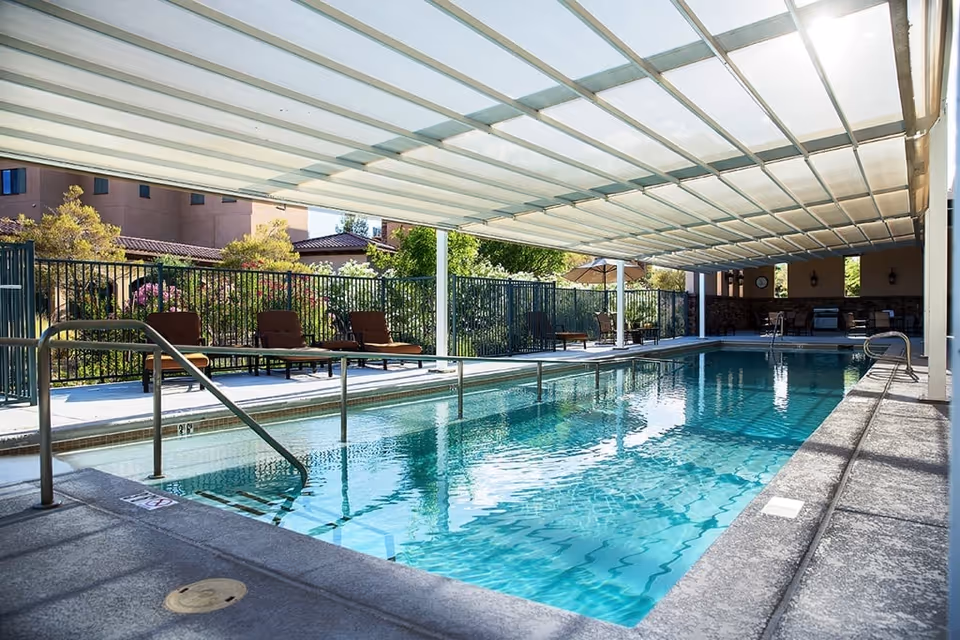 Covered outdoor swimming pool with clear blue water, surrounded by a concrete deck with metal handrails. Several lounge chairs and tables with umbrellas are placed along the fenced perimeter. Trees and buildings are visible in the background.