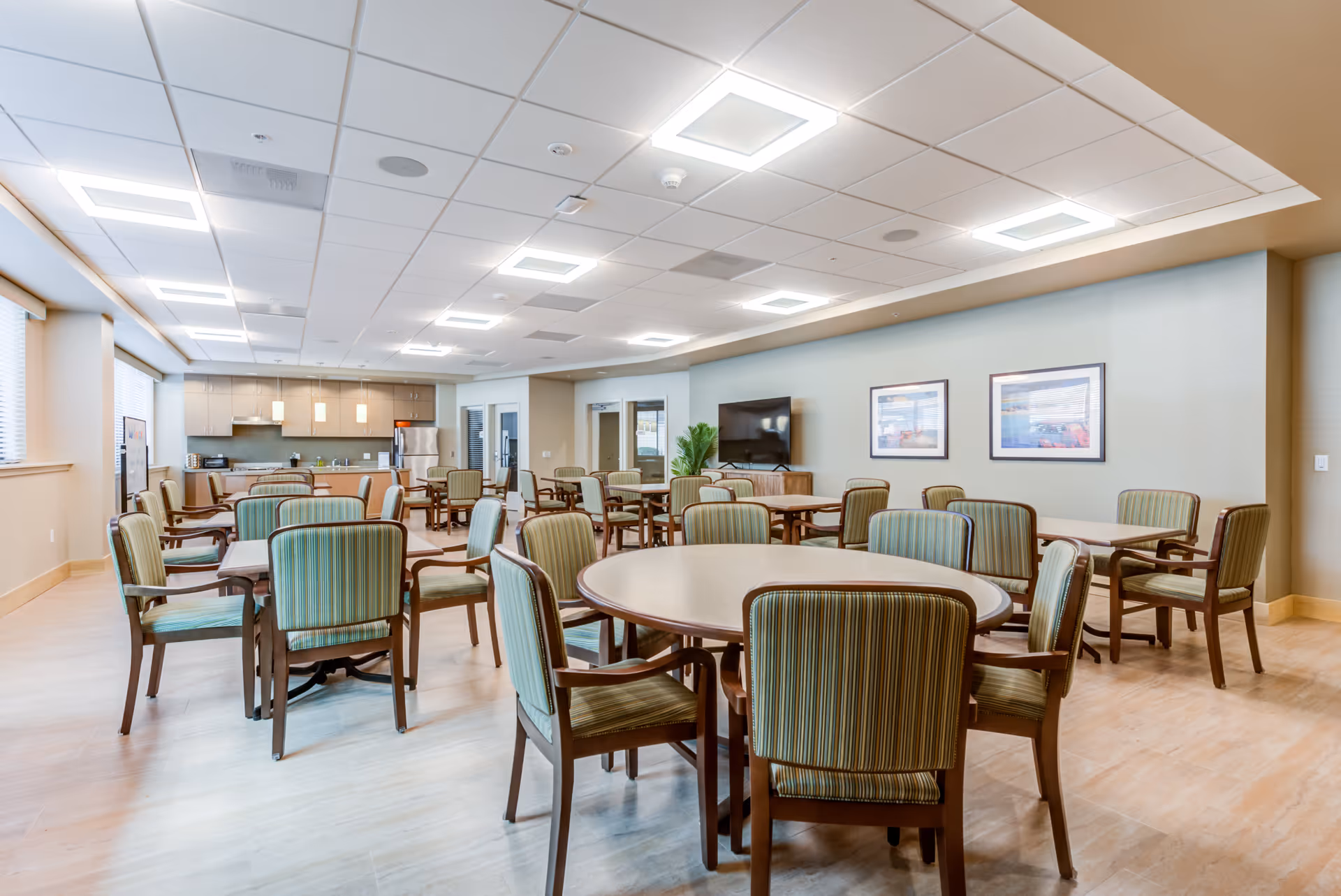 Spacious communal dining area with round and rectangular tables surrounded by green-striped chairs, a kitchenette and wall-mounted TV in the background.