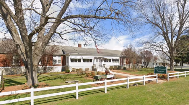 Exterior view of a single-story building with a white fence in front, leafless trees, a green lawn, and a sign that reads Alpha Concord Plantation. The building has a gray roof, white awnings over the windows, and an American flag near the entrance.