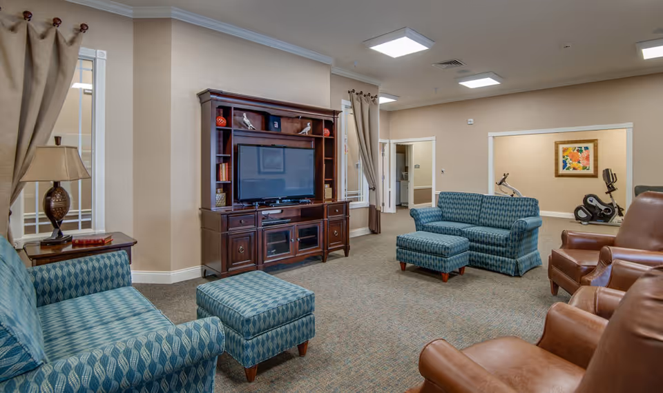 Comfortable common lounge with patterned sofas and ottomans, leather recliners, a large TV cabinet, and exercise equipment in the background.