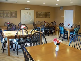 A dining room with multiple wooden tables and black chairs arranged neatly. Each table has a small vase with red and white flowers. The walls are painted beige and decorated with framed abstract artwork. There is a white door on the right side of the room.