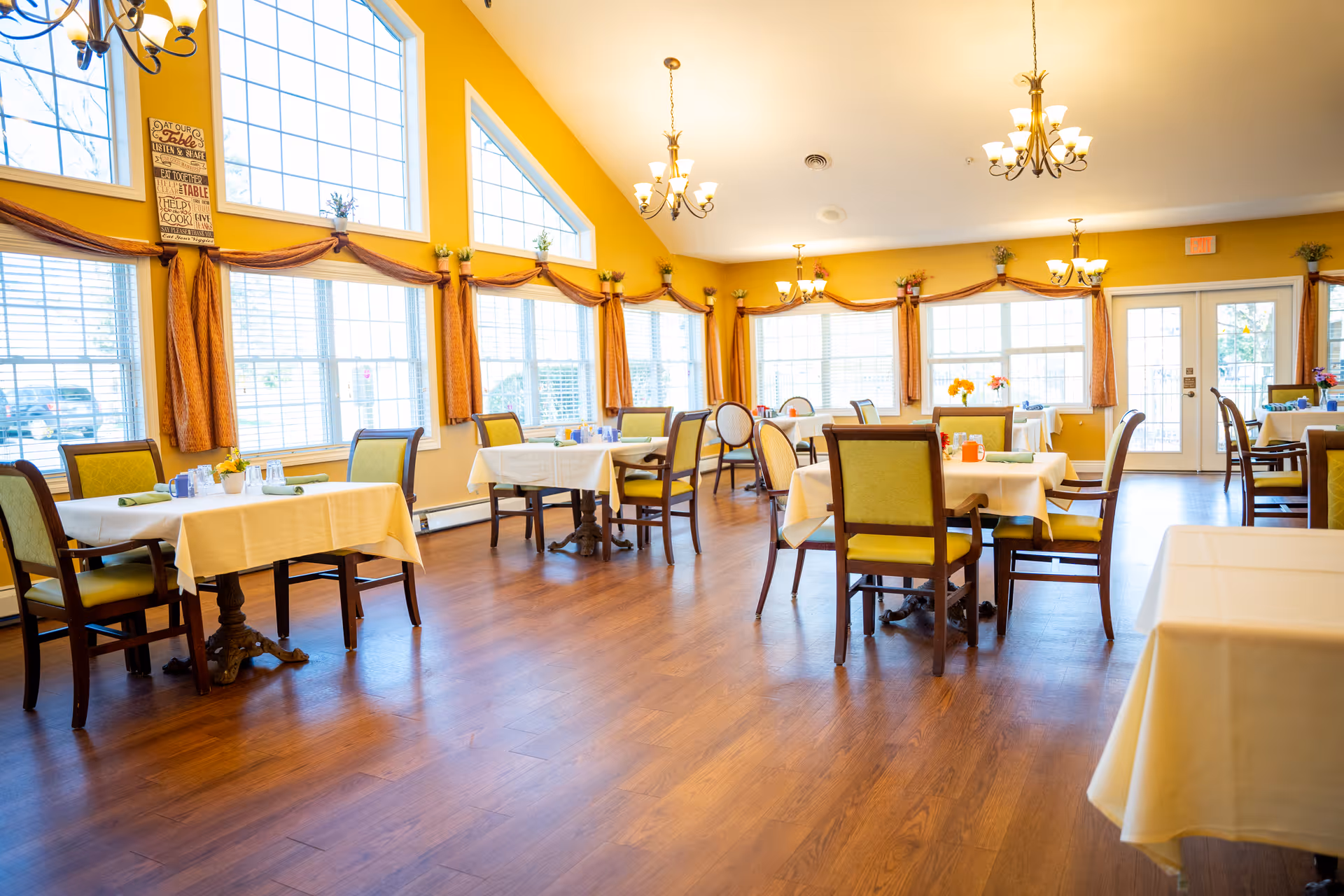 Bright and spacious dining room with multiple tables covered in white tablecloths, each surrounded by green cushioned chairs. Large windows with orange curtains allow natural light to fill the room. The walls are painted yellow, and chandeliers hang from the ceiling providing additional lighting. The floor is wooden, and there are decorative plants and flowers on some tables and window sills.