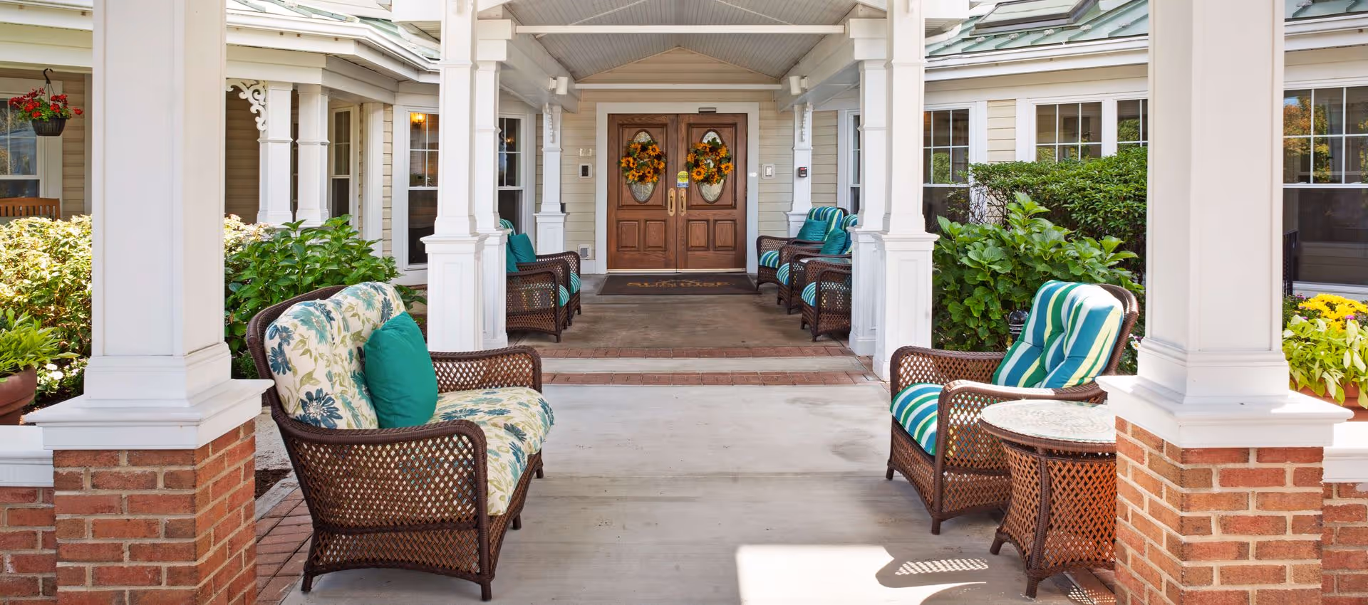 Covered outdoor seating area at the entrance of a building with wicker chairs and sofas featuring floral and striped cushions, surrounded by green shrubs and plants, with double wooden doors decorated with sunflower wreaths in the background.