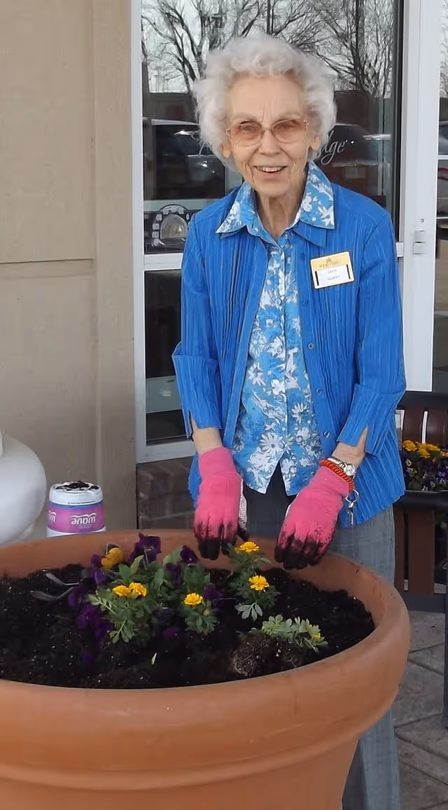 An elderly woman wearing pink gardening gloves, a blue floral shirt, and a blue jacket is planting flowers in a large terracotta pot outside a building with glass doors.