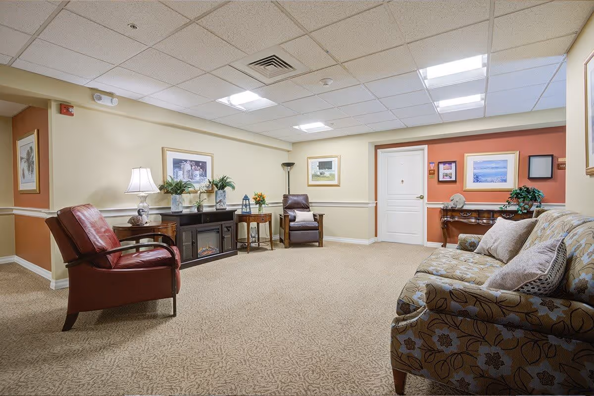 A cozy senior living facility common area with beige carpet and cream-colored walls accented by a warm orange section. The room features a floral-patterned sofa with pillows, two leather armchairs, a wooden side table with a lamp, a black cabinet with decorative plants and lanterns, and framed artwork on the walls. A white door and a wooden console table with more decor are visible in the background.
