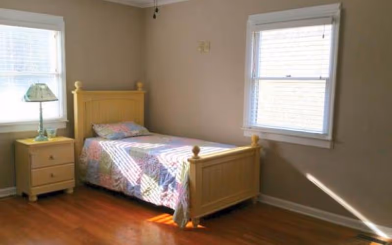 A small bedroom with a single wooden bed featuring a colorful quilt. Next to the bed is a wooden nightstand with a decorative lamp. The room has two windows with white blinds allowing natural light to enter, and the floor is wooden.