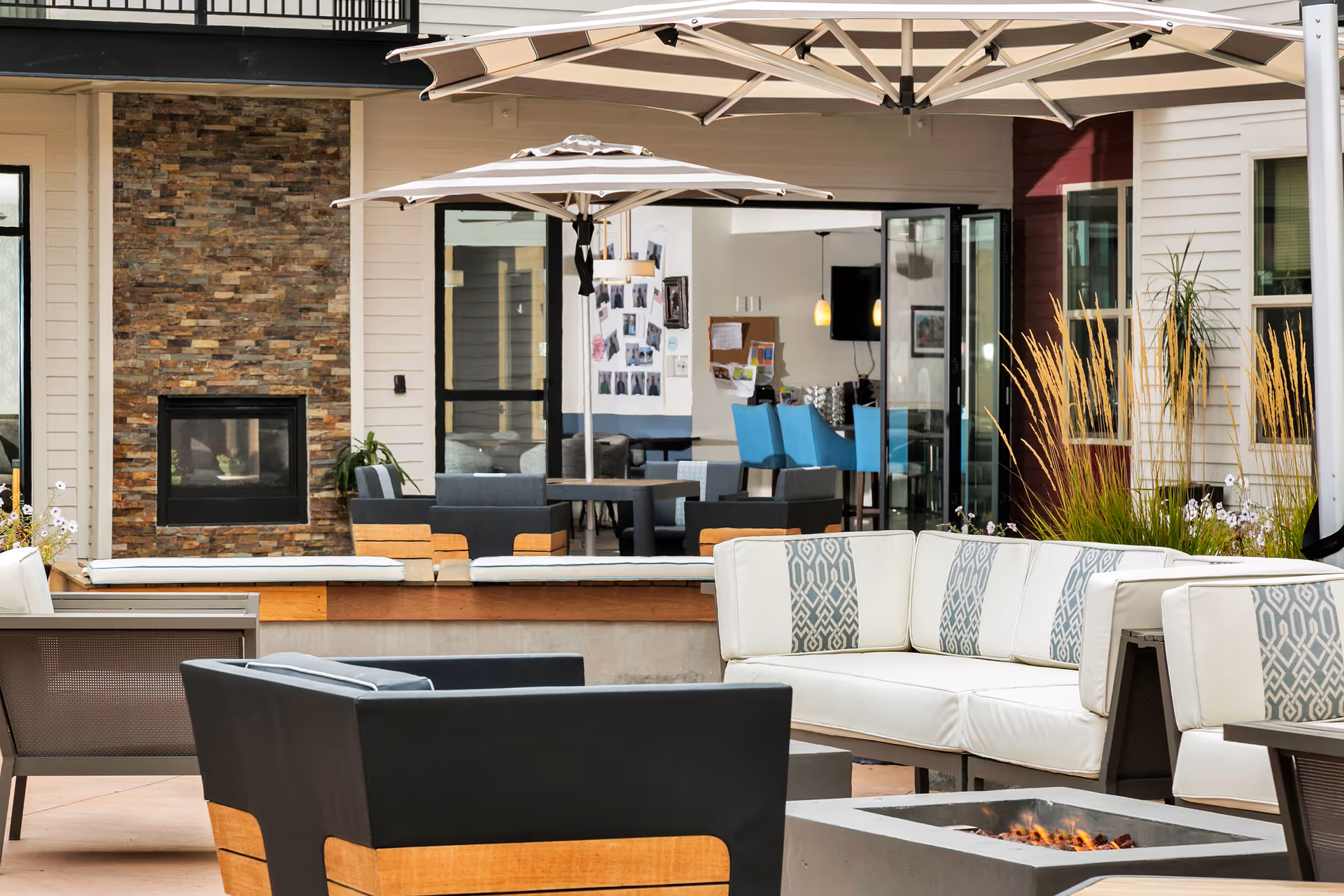 Outdoor patio with cushioned lounge seating, umbrellas, a fire pit, and a stone-clad fireplace in front of the facility.