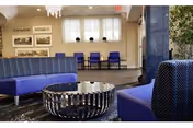 A modern and stylish common area with purple cushioned seating, a round metallic coffee table, framed pictures on the wall, and large windows with white curtains letting in natural light.