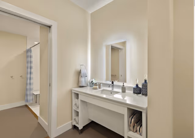 Bright bathroom with a white vanity, illuminated mirror, sink and towel shelving, and an open doorway revealing a shower with a blue checkered curtain.