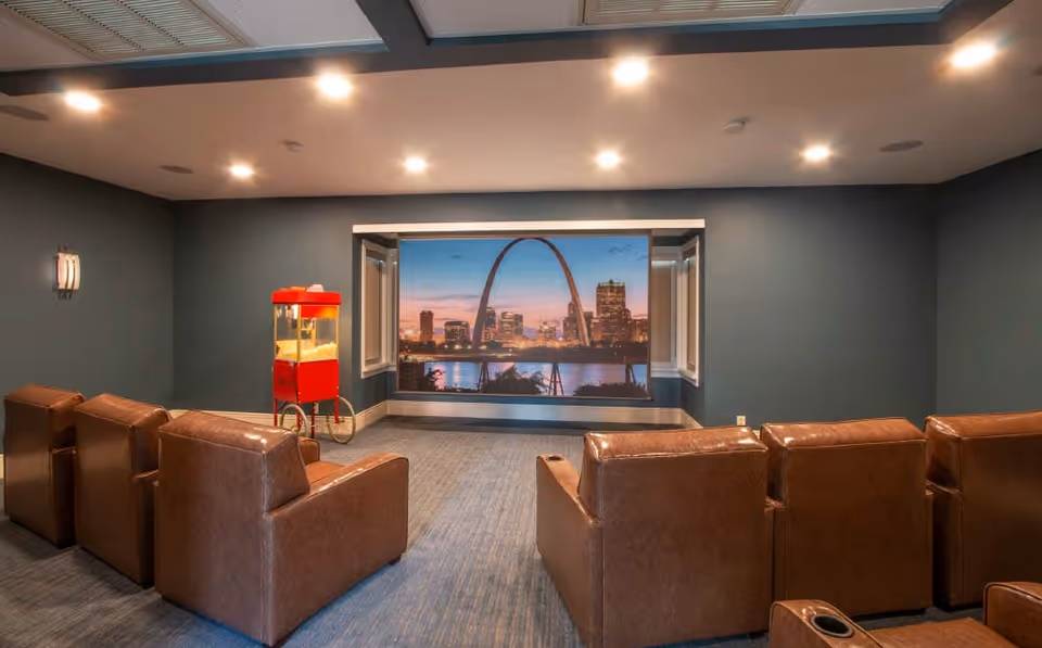 A small theater room with six brown leather recliners arranged in two rows facing a large screen displaying a cityscape with a prominent arch. A red popcorn machine is positioned to the left side of the screen. The room has dark walls and ceiling lights.