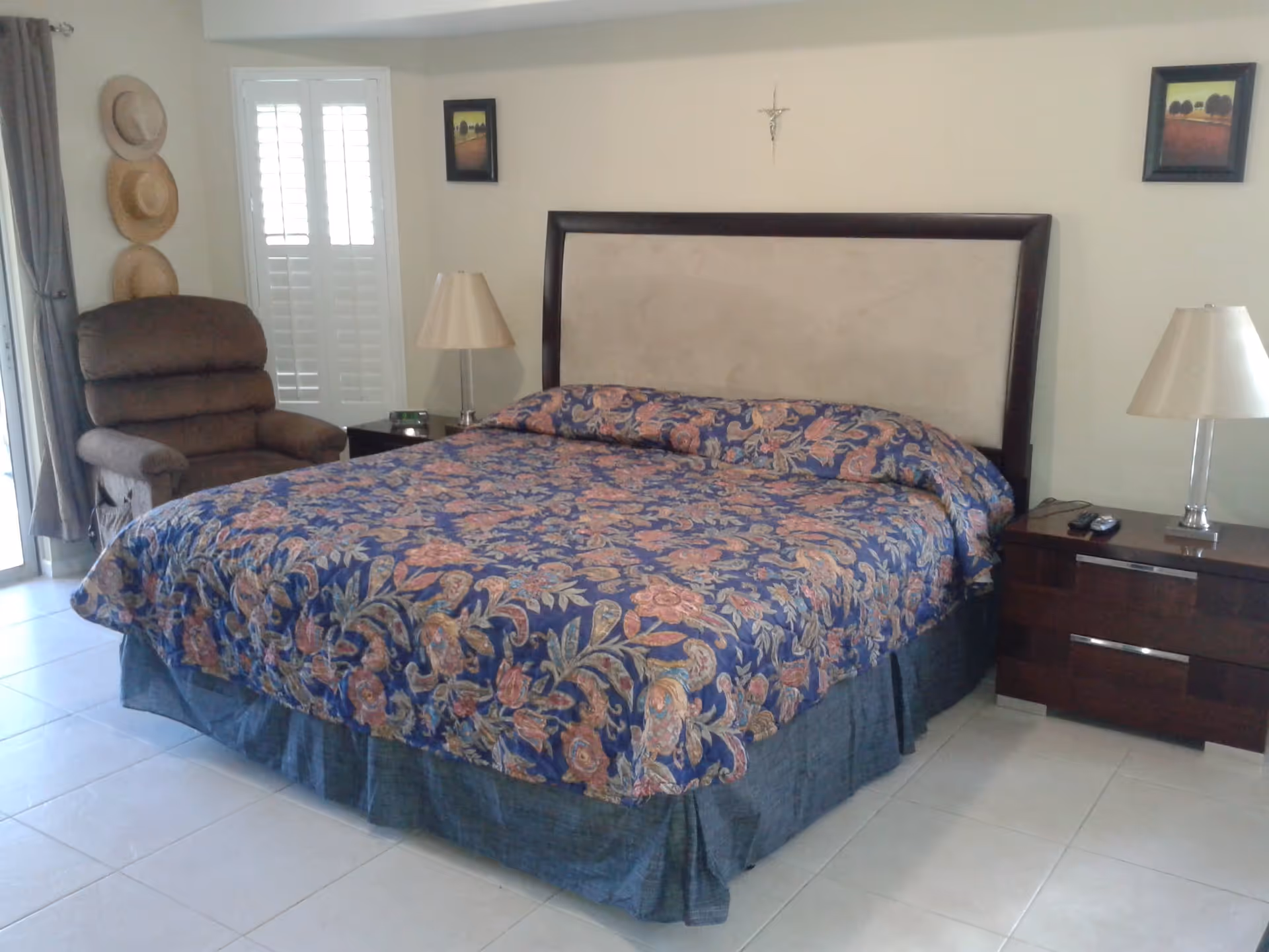 A bedroom with a large bed covered in a floral patterned bedspread. There are two nightstands on either side of the bed, each with a lamp. A recliner chair is positioned near a window with white shutters and curtains. Three hats hang on the wall to the left of the window, and two framed pictures and a crucifix are mounted on the wall above the bed.