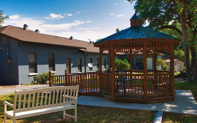 Outdoor area of Lakeland Manor Assisted Living Facility featuring a wooden gazebo with a green roof, surrounded by a wooden railing and a concrete pathway. A white bench is placed on the grass nearby, with a gray building and trees in the background under a partly cloudy sky.