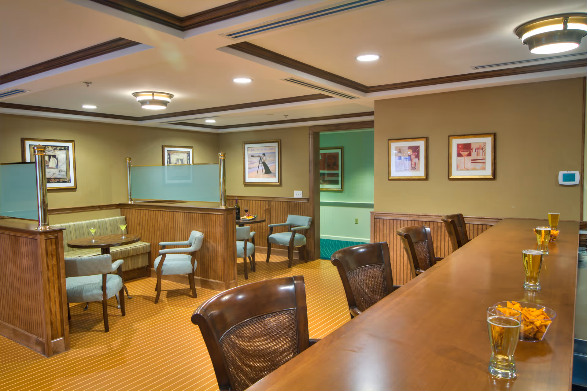 Interior view of a senior living facility lounge area with a wooden bar counter on the right side, featuring several glasses of beer and bowls of snacks. On the left side, there are small seating areas with cushioned chairs and tables, separated by wooden partitions with frosted glass panels. The walls are decorated with framed artwork, and the ceiling has recessed lighting and decorative light fixtures.