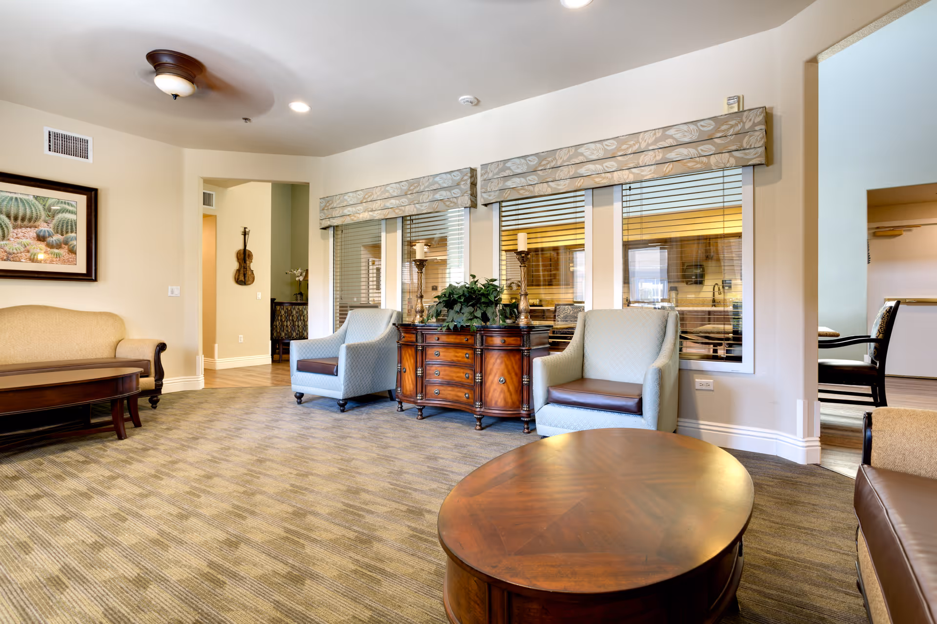 Bright communal living room with armchairs, a sofa, wooden coffee table and sideboard in front of windows with blinds.