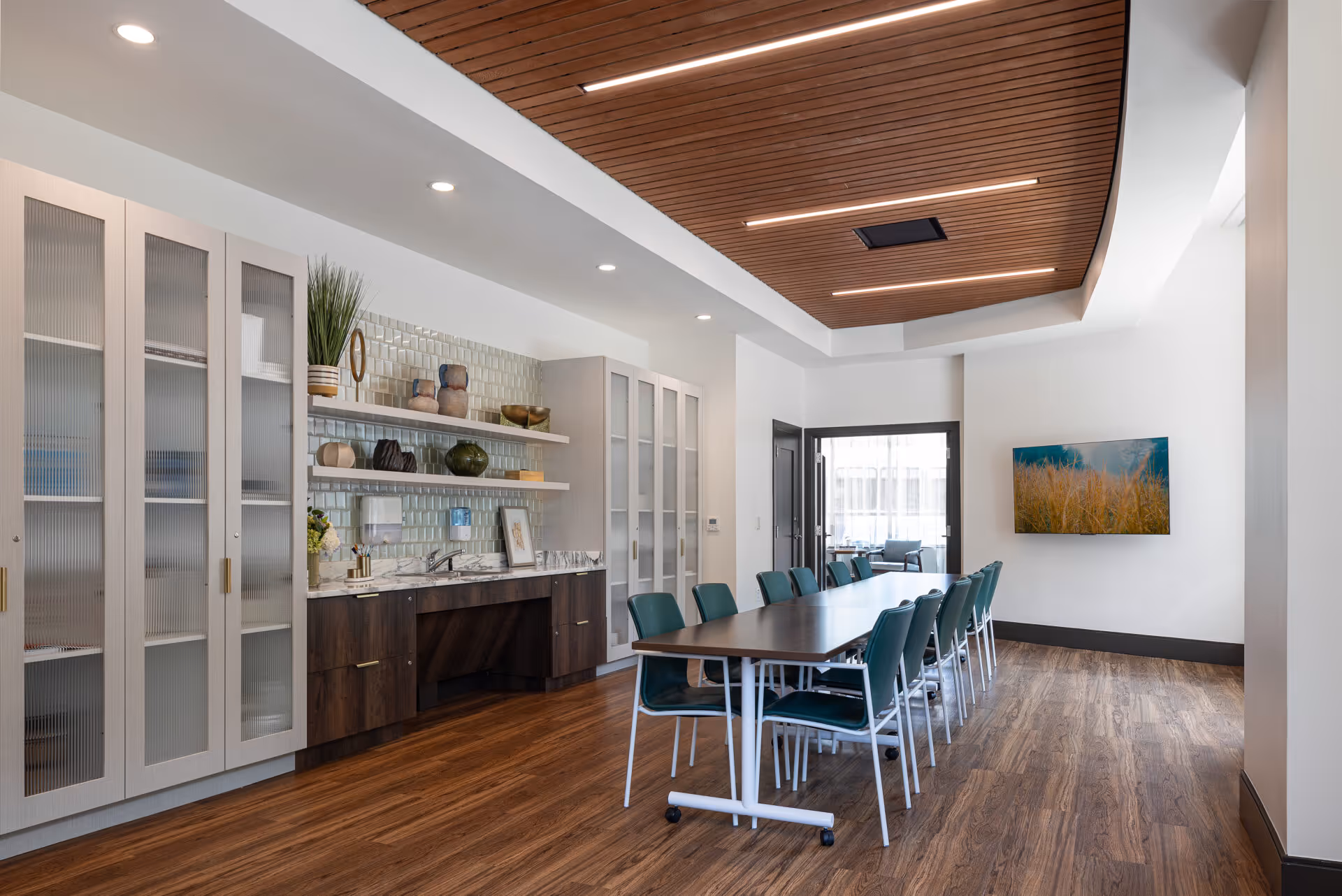 A modern conference or dining room with a long rectangular table surrounded by green chairs. The room features wood flooring, a wooden slatted ceiling with recessed lighting, and a wall-mounted TV displaying an image of tall grass. Along one wall, there are tall cabinets with frosted glass doors, open shelves with decorative items, and a countertop with a sink.