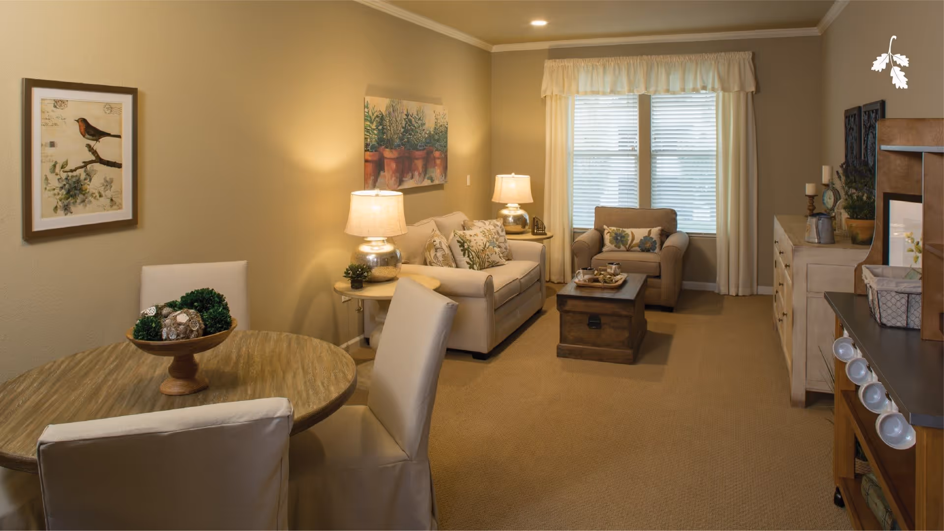 A cozy living room and dining area with beige walls and carpet. The dining area has a round wooden table with four white cushioned chairs and a decorative centerpiece. The living room features a beige sofa with floral pillows, a matching armchair, a wooden coffee table, two table lamps, and a window with white curtains. Wall art and decorative items are displayed on the walls and furniture.