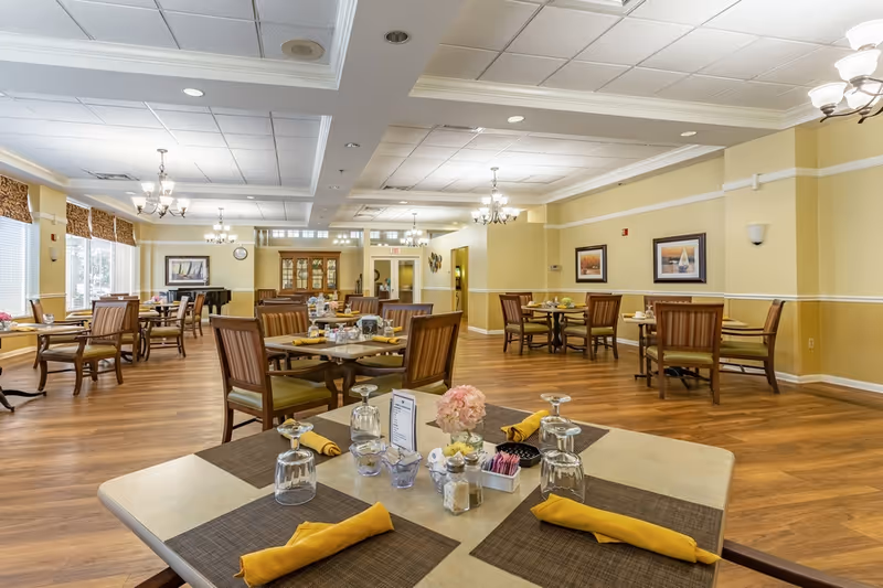 A spacious dining room in a senior living facility with multiple wooden tables and chairs arranged neatly. Each table is set with placemats, yellow napkins, upside-down glasses, and small decorative items including a flower centerpiece. The room has large windows with patterned valances, wooden flooring, soft yellow walls, and several chandeliers providing warm lighting. Framed artwork and a china cabinet are visible in the background.