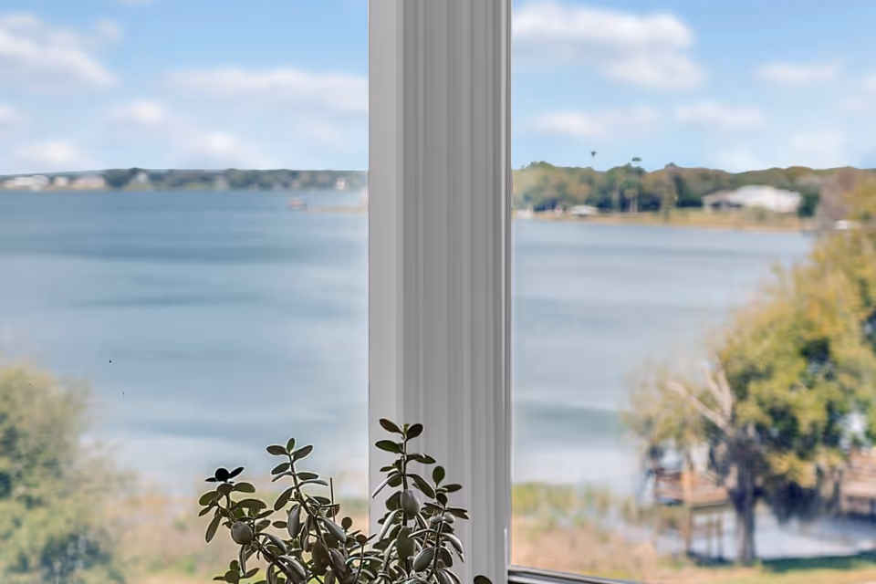 View of a large body of water and distant shoreline seen through a window with a small potted plant on the windowsill.