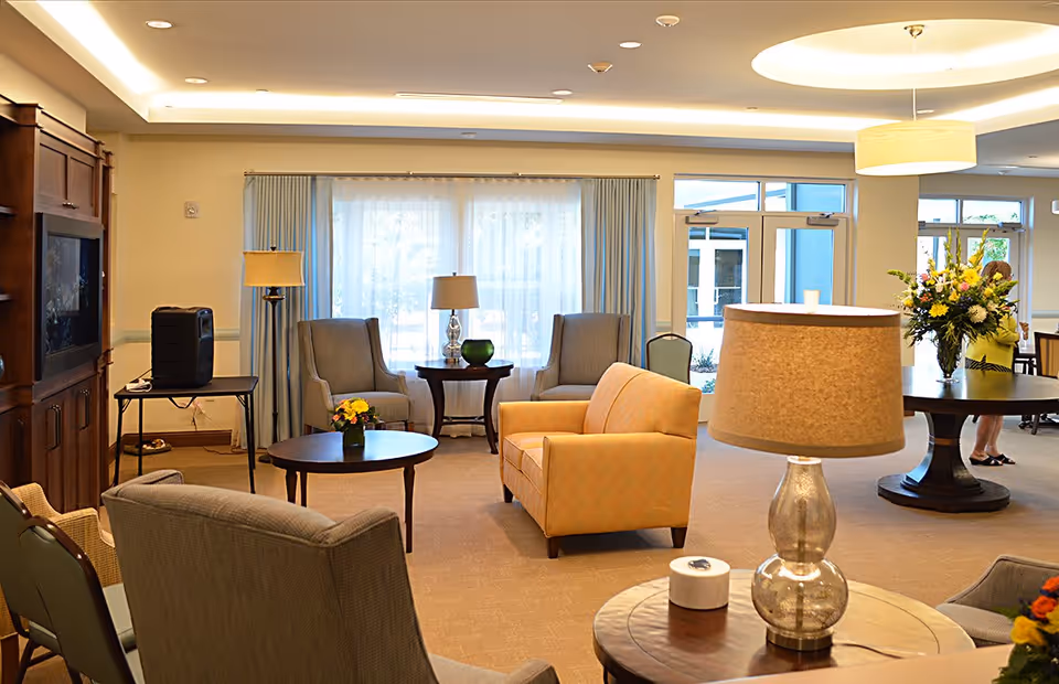 A well-lit living room area in Paradise Valley Estates featuring comfortable seating including armchairs and a loveseat, wooden tables with decorative lamps and flower arrangements, large windows with sheer curtains, and a wooden entertainment center. A woman is seen in the background near a round table with a large floral arrangement.