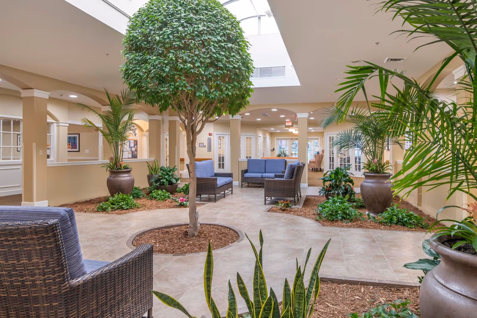 Atrium-style indoor lounge with wicker seating, large potted plants, and a central tree beneath a skylight.