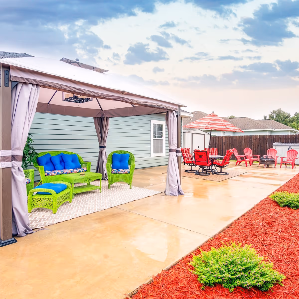 Outdoor patio area with a covered gazebo featuring green wicker furniture with blue cushions, a concrete floor, a red and white striped umbrella over a table with red chairs, additional red chairs around a fire pit, and landscaping with red mulch and green shrubs.