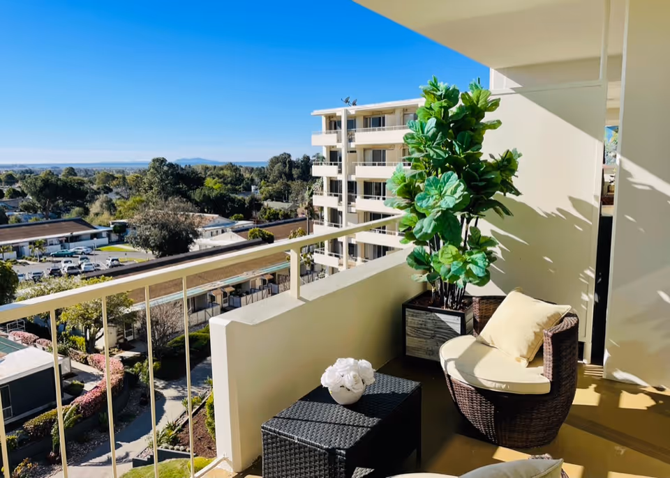 Balcony with a wicker chair with cream cushions, a small black wicker table with a white flower arrangement, and a large potted green plant. The balcony overlooks a residential area with trees, buildings, and a clear blue sky.