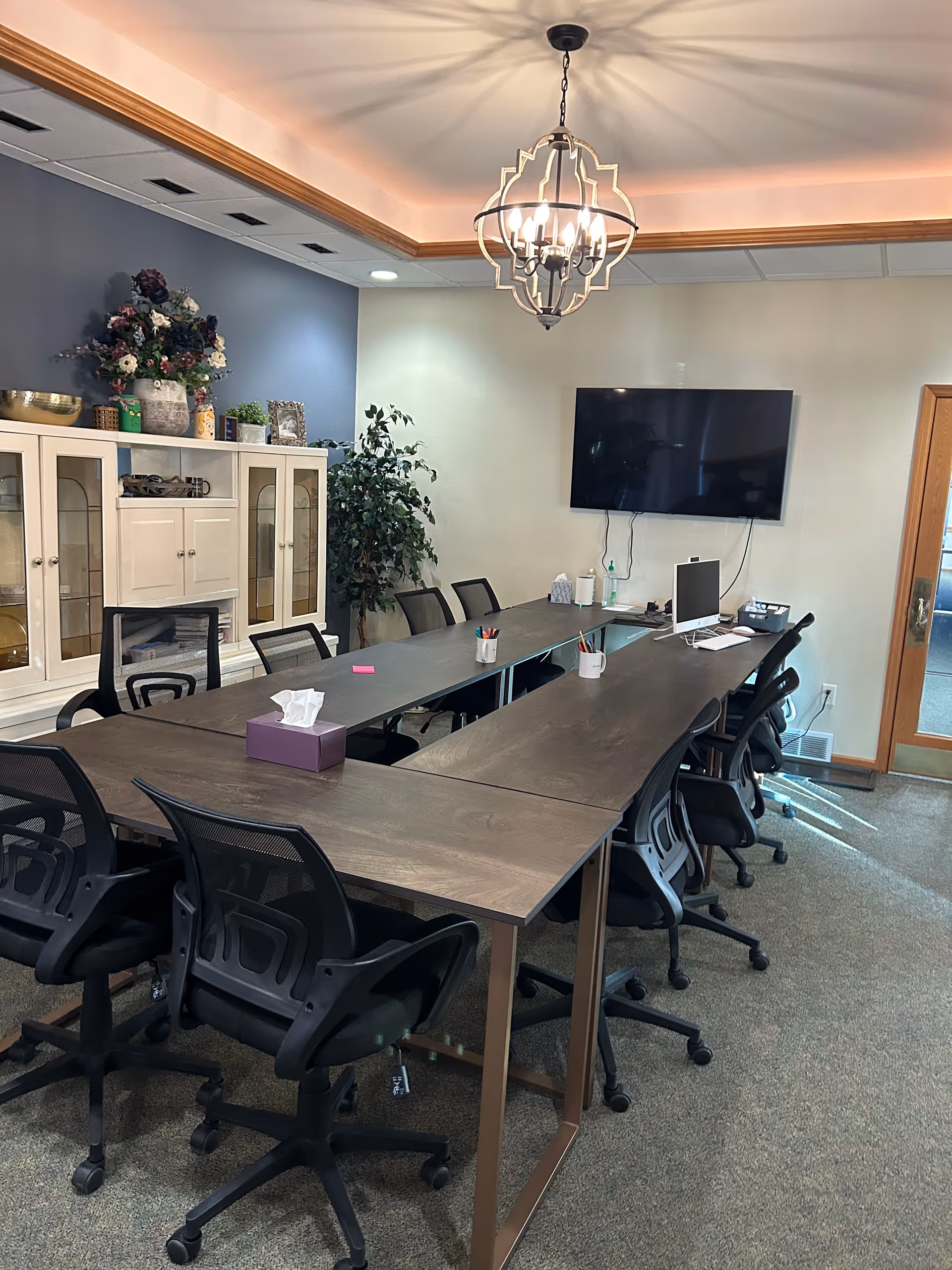 A conference-style meeting room with long tables and rolling office chairs, a wall-mounted TV, decorative cabinet, plants, and a chandelier.