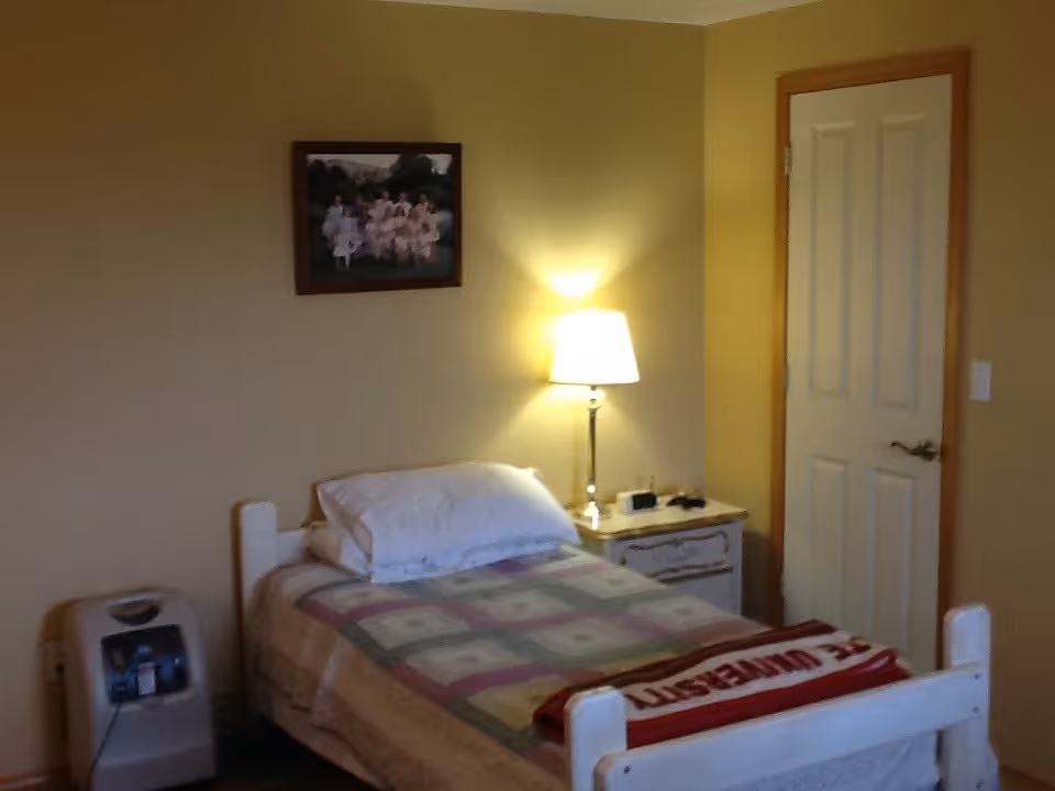 A small bedroom with a single bed covered with a patterned quilt and a pillow. Next to the bed is a nightstand with a lamp, a clock, and some small items. There is a door on the right side of the room and a framed picture hanging on the wall above the bed. An oxygen concentrator is placed on the floor to the left of the bed.