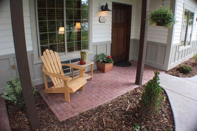 A small outdoor porch area with a wooden Adirondack chair and a small side table holding a potted plant and a red cup. The porch has brick flooring and is adjacent to a house with white and gray siding, a wooden door, and a window with blinds. There are plants and mulch surrounding the porch area.