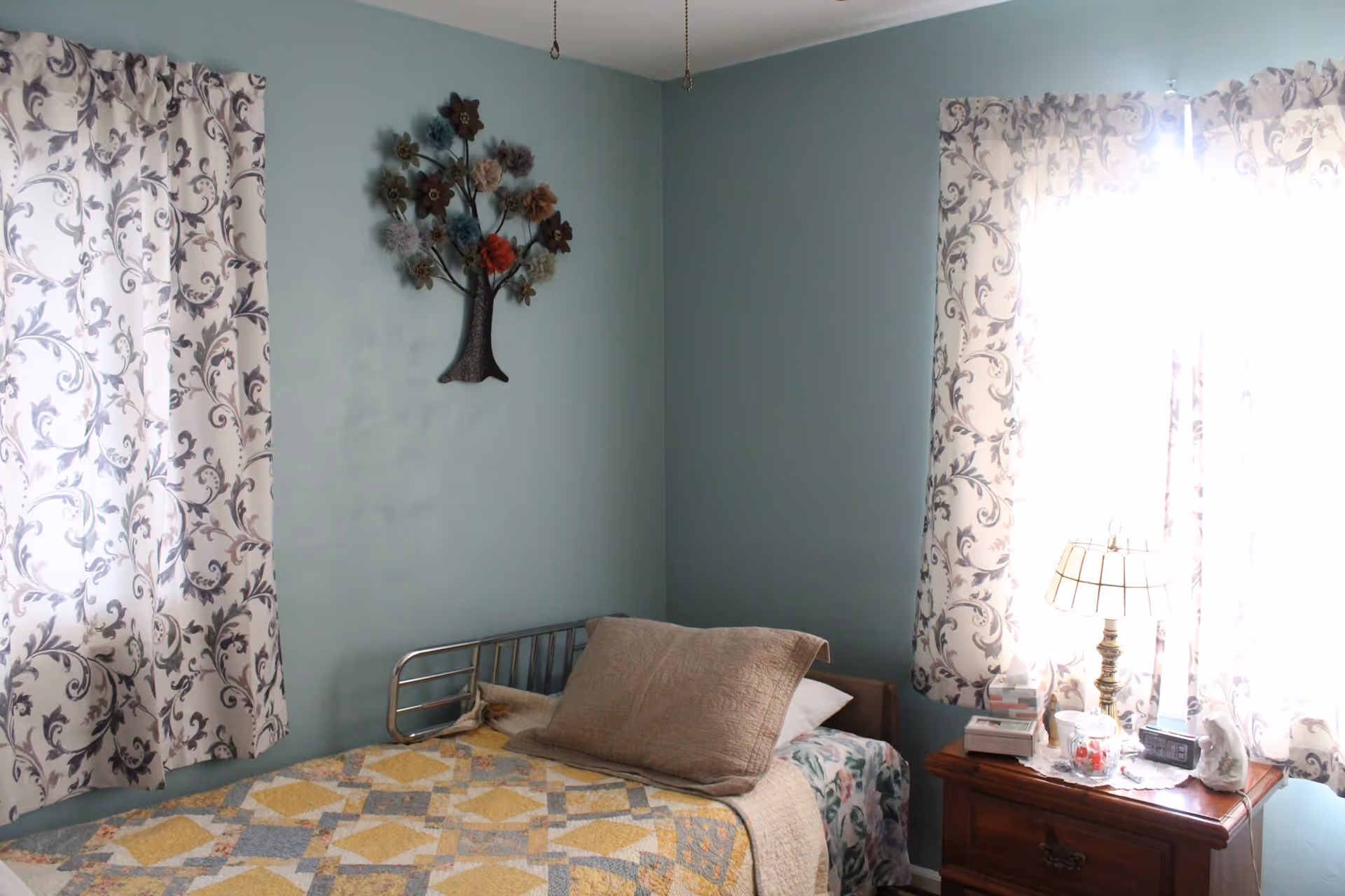A small bedroom with light blue walls, a single bed with a yellow and blue patchwork quilt, and a beige pillow. There are two windows with white and gray patterned curtains letting in bright light. A wooden nightstand next to the bed holds a lamp with a stained glass shade, a tissue box, a clock, and some decorative items. A metal wall decoration shaped like a tree with colorful flowers is mounted on the wall above the bed.