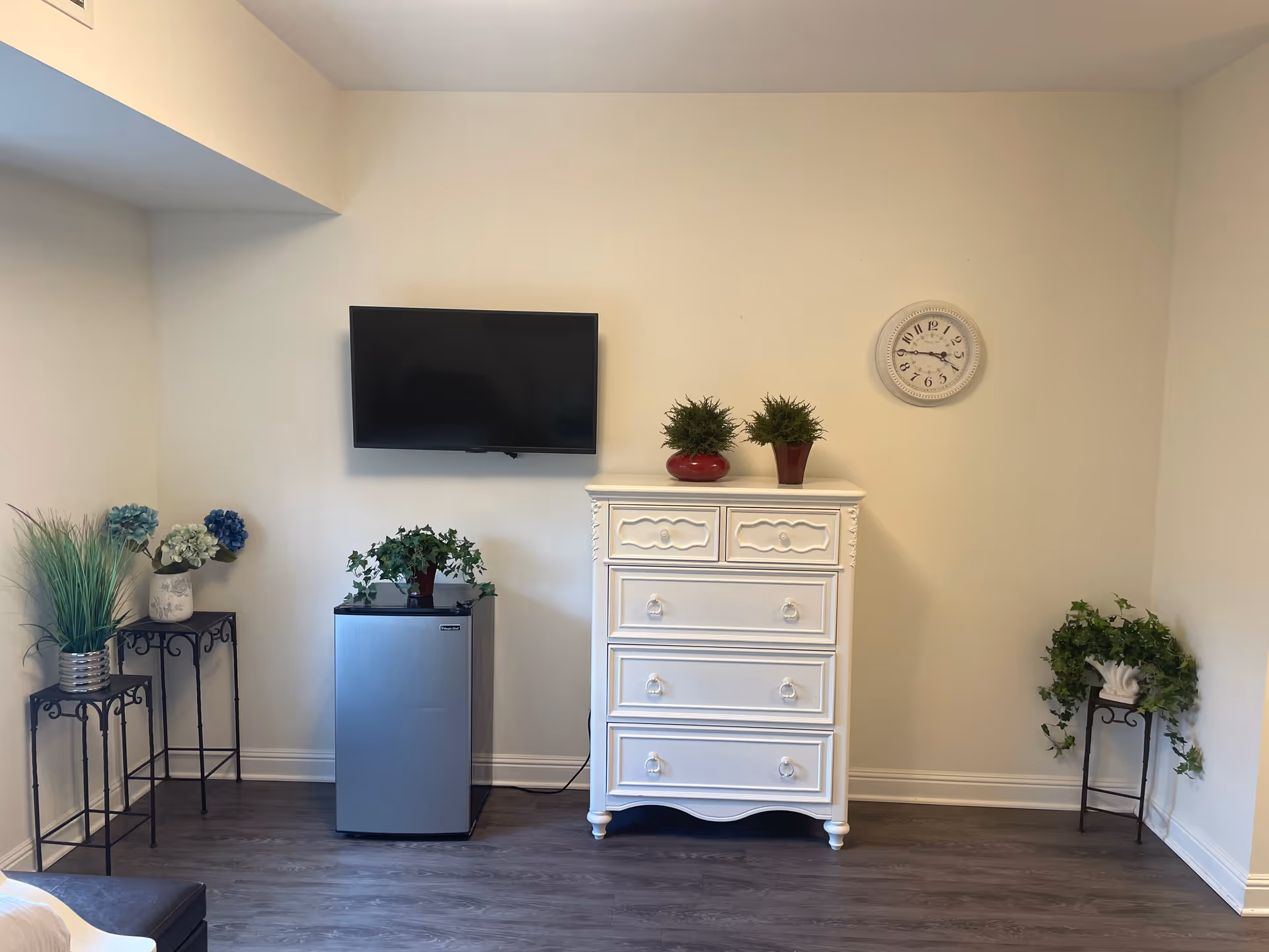 Wall-mounted TV above a small refrigerator and a white dresser decorated with potted plants and a wall clock in a simple interior room.