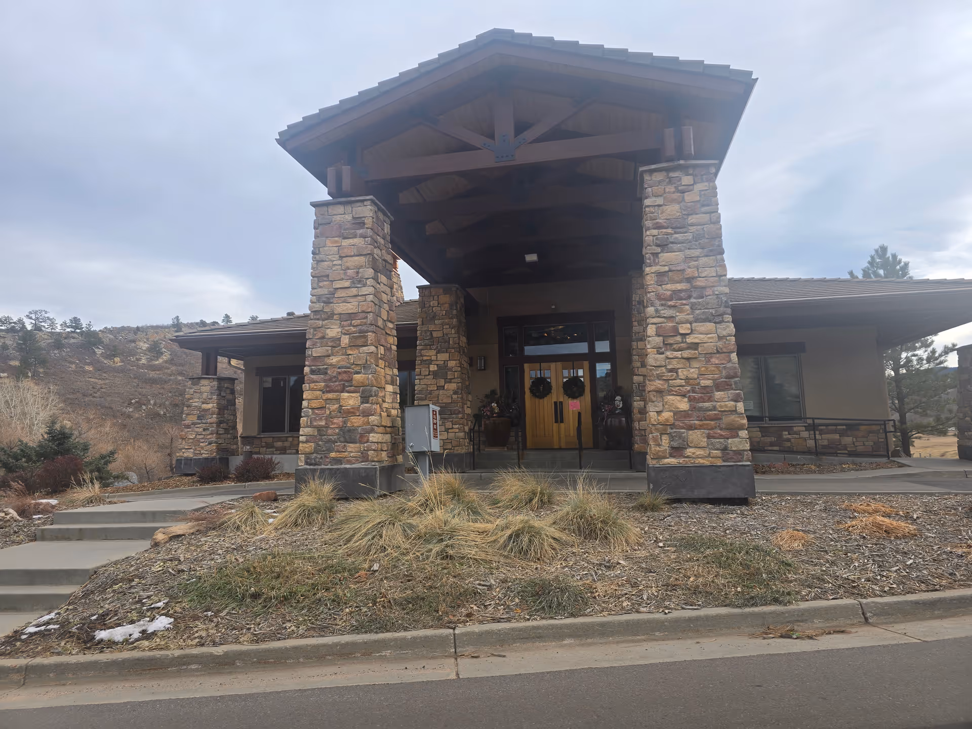 Front exterior view of a building with stone pillars and a covered entrance. The building has wooden double doors decorated with wreaths and is surrounded by dry grass and shrubs. The sky is overcast.