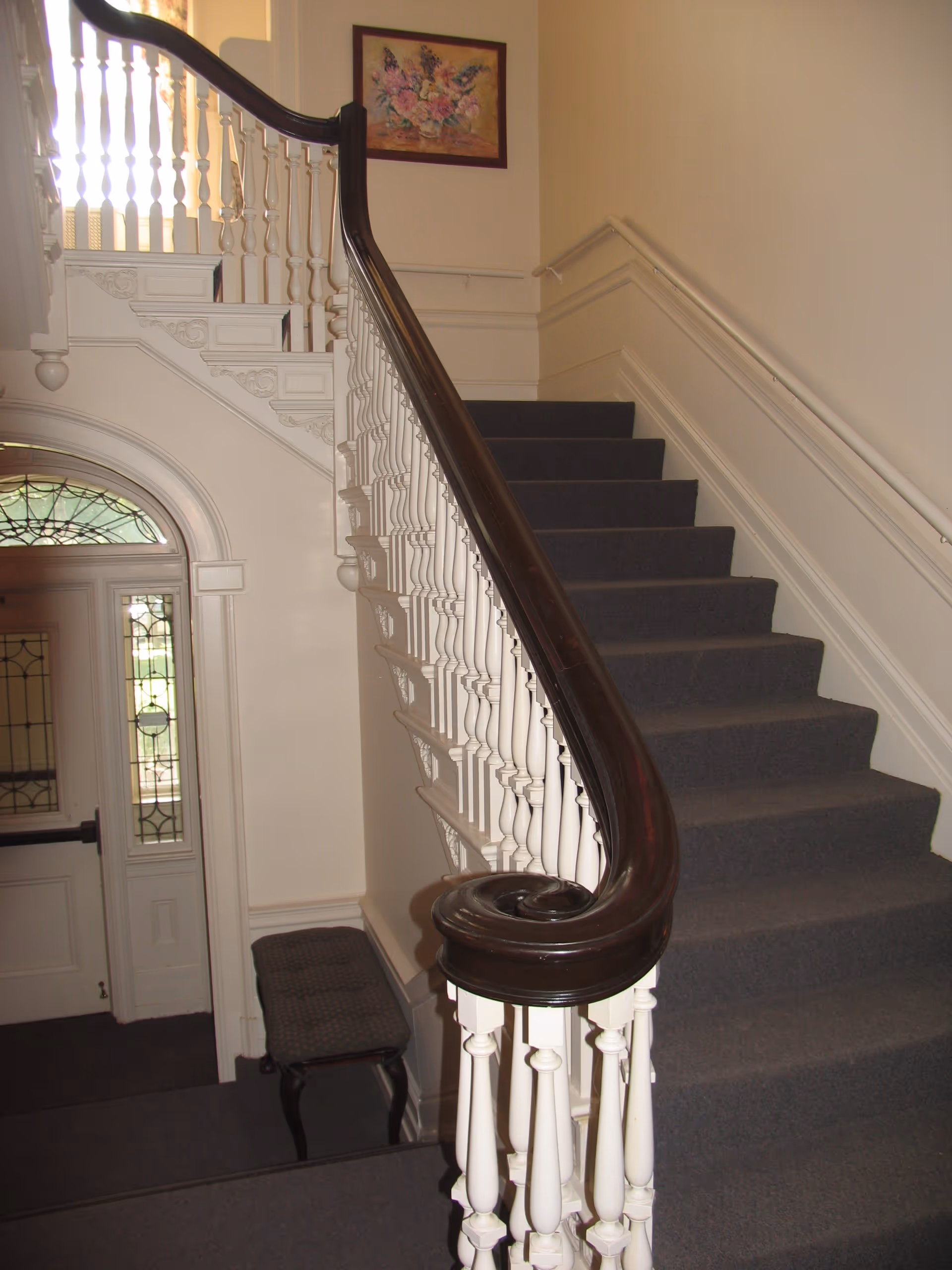 Interior view of a staircase with a wooden handrail and carpeted steps, featuring a decorative painting on the wall and a bench at the bottom.