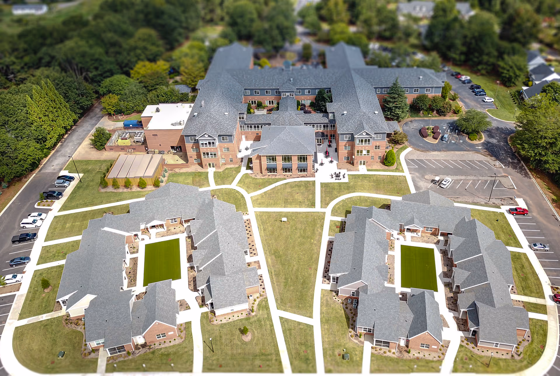 Aerial view of Rosecrest Retirement Community showing multiple large buildings with gray roofs arranged around green lawns and walkways, surrounded by trees and parking areas.