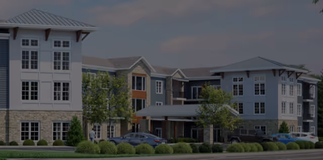 Three-story senior living building with stone and siding facade, landscaped entrance and parked cars.