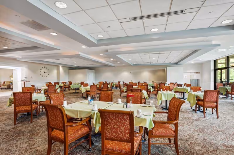 A spacious dining room with multiple tables covered in light green tablecloths, each set with white cups and silverware. The chairs have wooden frames with red patterned upholstery. The room has a carpeted floor, a white ceiling with recessed lighting, and large windows on the right side letting in natural light.