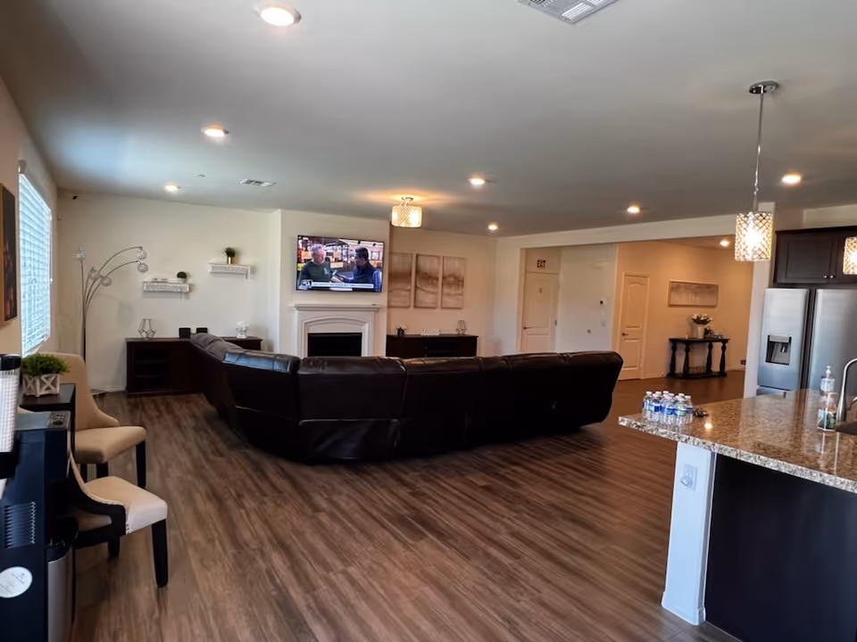 A spacious living room area with a large dark brown leather sectional sofa facing a wall-mounted TV above a white fireplace. The room has wood flooring, recessed ceiling lights, and light-colored walls. To the right, there is a kitchen area with granite countertops, pendant lights, a stainless steel refrigerator, and several water bottles on the counter. On the left side, there are chairs and a water dispenser near a window with blinds. Decorative shelves and artwork are on the walls.
