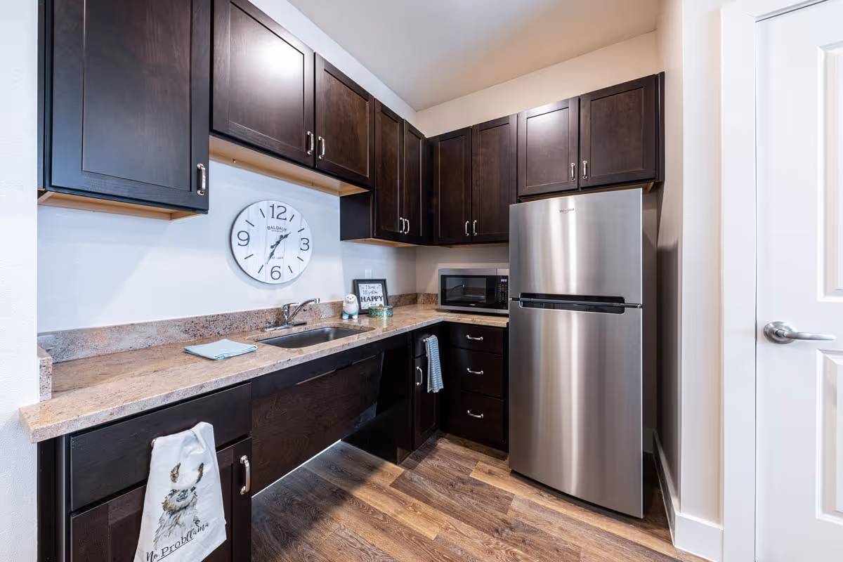 Small modern kitchenette with dark wood cabinets, a stainless-steel refrigerator, sink, microwave, wall clock, and wood-look flooring.