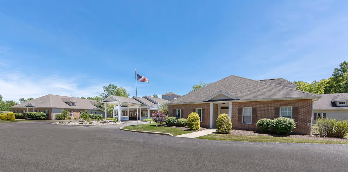 Single-story brick assisted living building with an entrance canopy, American flag, landscaped shrubs and a paved driveway under a clear blue sky.