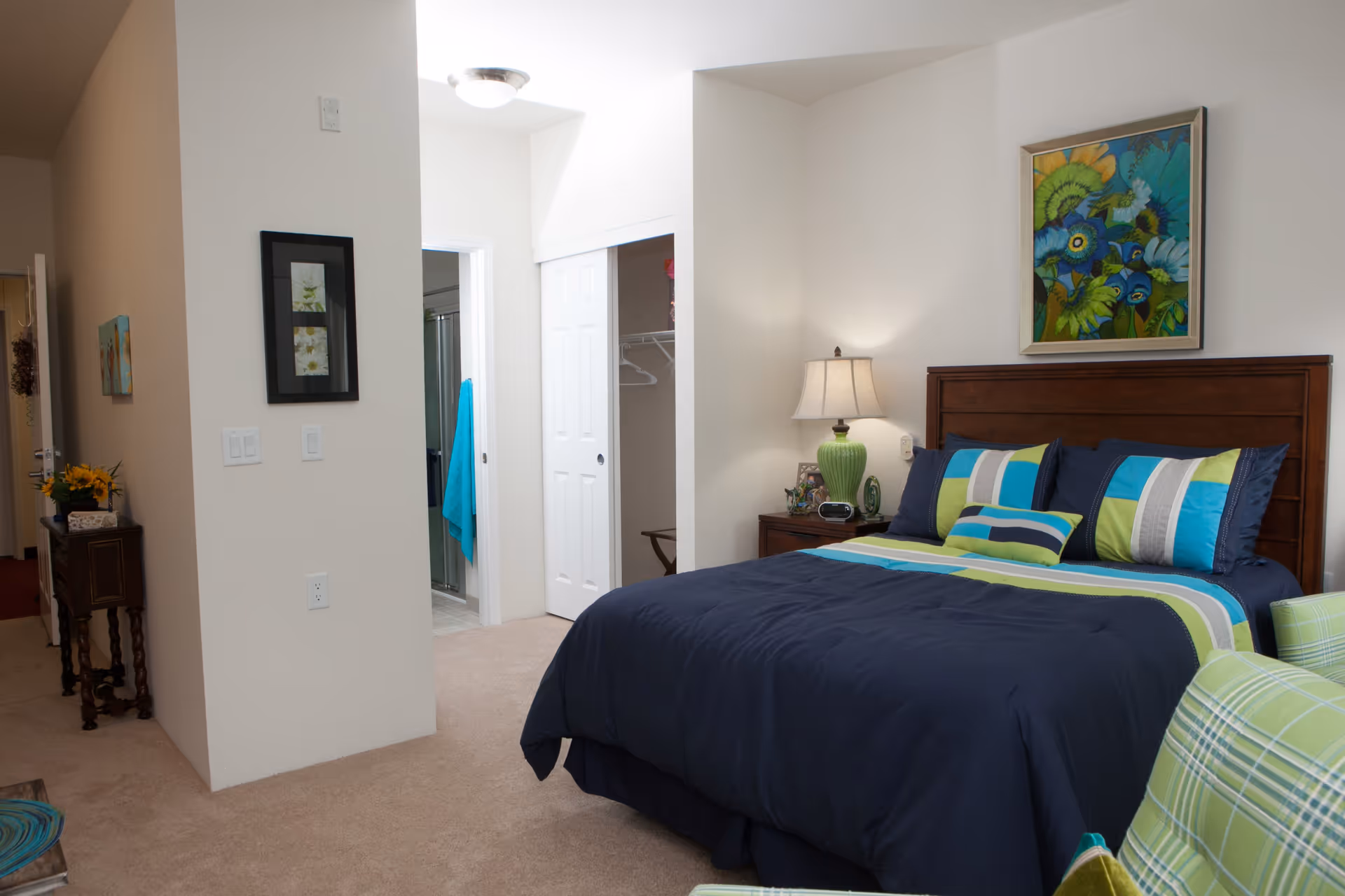 A bedroom in a senior living facility with a neatly made bed featuring blue, green, and white striped bedding. There is a wooden headboard, a nightstand with a green lamp, and a colorful floral painting on the wall above the bed. To the left, there is an open closet with white sliding doors and a bathroom entrance with a blue towel hanging inside. The room has beige carpet and light-colored walls.