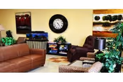 A cozy living room area with a brown leather sofa, a purple armchair, a patterned ottoman, a small table with a lamp, and a fish tank on a stand. The walls are decorated with a large round clock and two colorful paintings. There are also several green plants adding a touch of nature to the space.