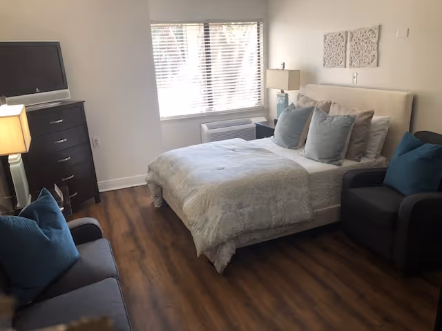 A cozy bedroom with a bed covered in light-colored bedding and multiple pillows. There is a dark armchair with a blue cushion to the right of the bed and a dark sofa with blue cushions to the left. A window with blinds lets in natural light, and a dresser with a TV on top is positioned against the wall. The room has wooden flooring and light-colored walls with decorative wall art above the bed.