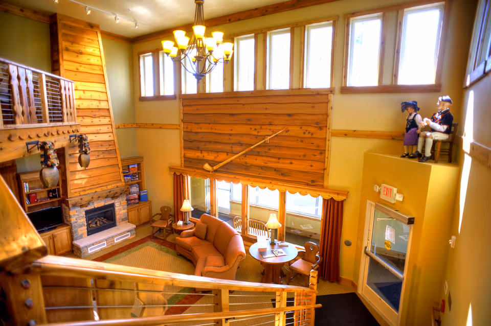 Sunlit two-story common lounge with wooden walls and railings, a stone fireplace, sofa and table seating under tall windows.