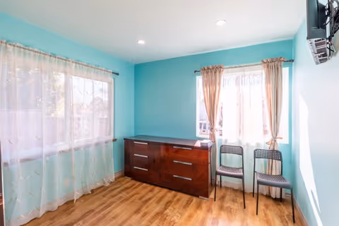 A bright room with light blue walls and wooden flooring featuring two windows with sheer curtains. There is a dark wooden dresser with multiple drawers against one wall and two black chairs placed near one of the windows. A flat-screen TV is mounted on the wall to the right.